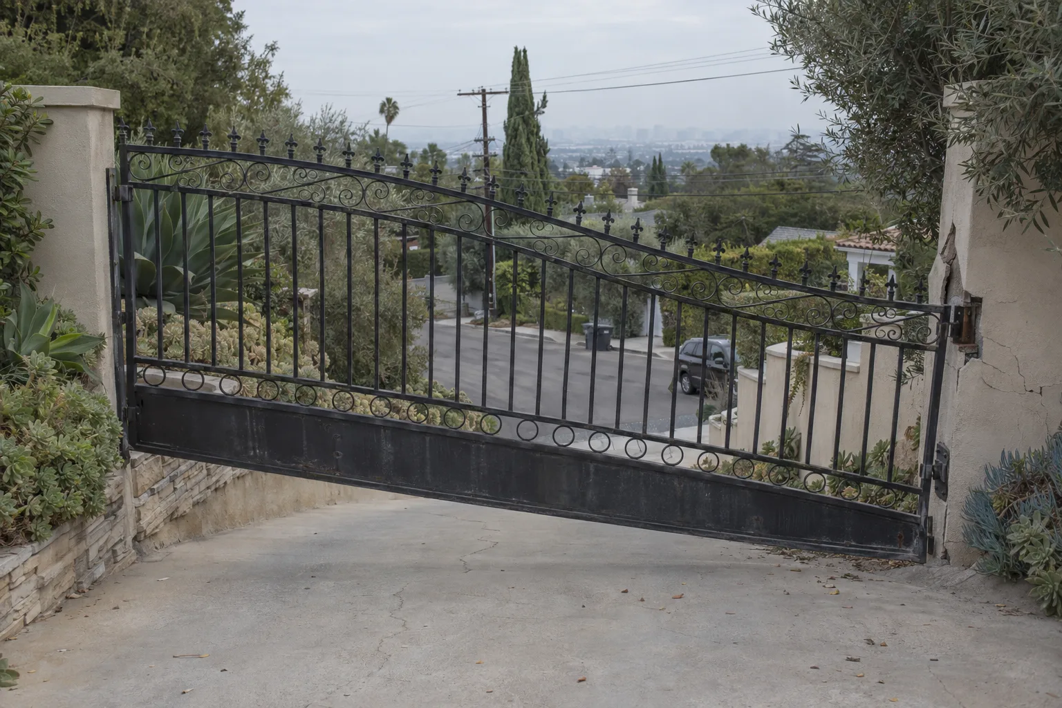 Ornamental iron entry gate sagging at the far corner beside a broken hinge, on a sloped Los Angeles driveway, daylight